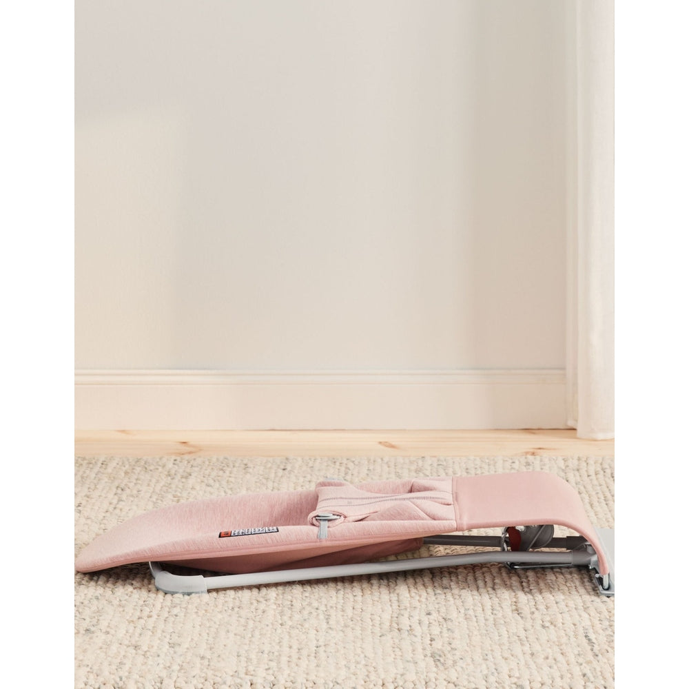 BabyBjorn Bouncer Bliss in soft pink, folded flat on a beige rug with its lightweight frame exposed.