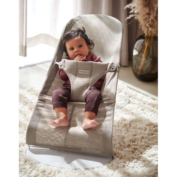 BabyBjorn Bouncer Bliss gray mesh infant bouncer; baby sits securely with feet dangling in a cozy living room.