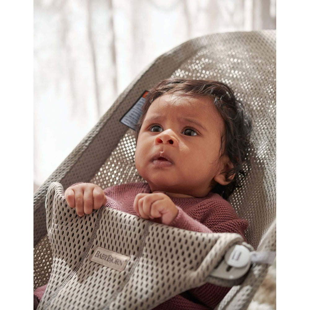 BabyBjorn Bouncer Bliss in grey mesh, infant seated and gripping the front edge in a curious moment.