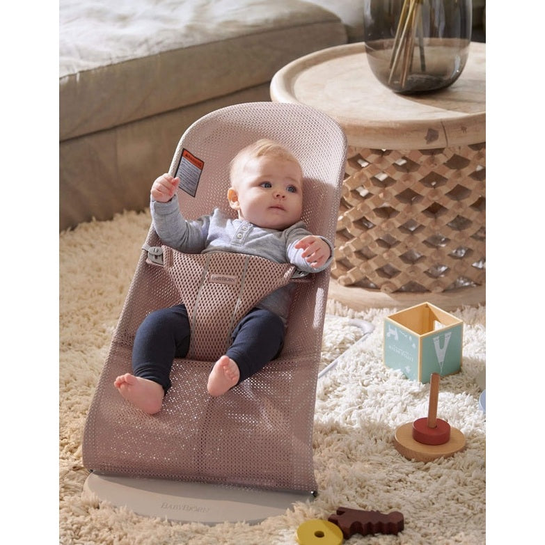 BabyBjorn Bouncer Bliss in pink mesh, baby strapped in and reaching hands up in a cozy living room.