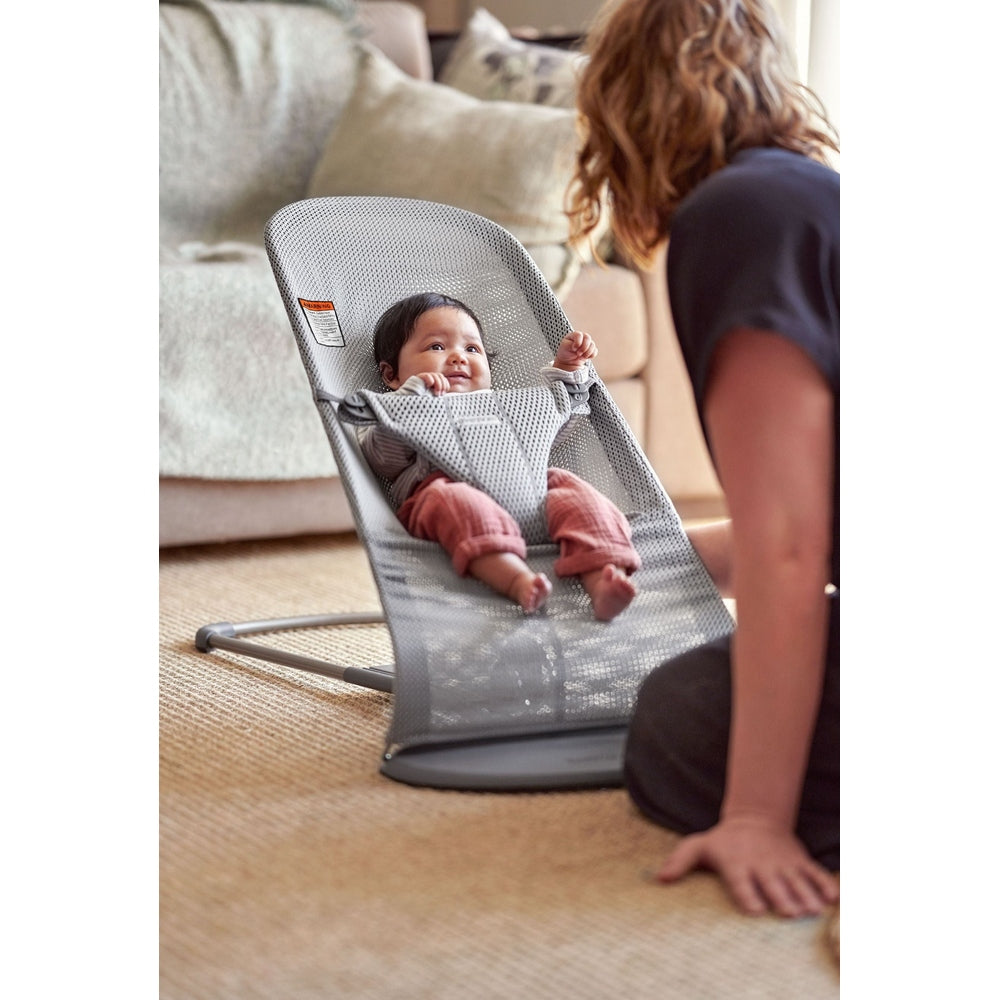 BabyBjorn Bouncer Bliss gray mesh infant bouncer in living room, baby smiling and kicking as caregiver watches.