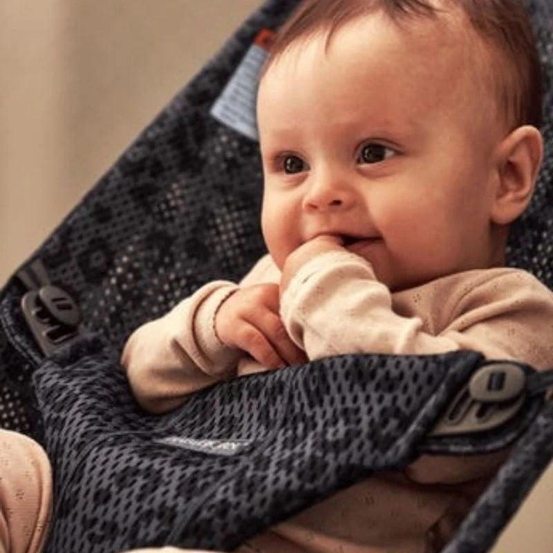 BabyBjorn Bouncer Bliss in dark grey mesh, baby calmly smiles and sucks fingers while seated.