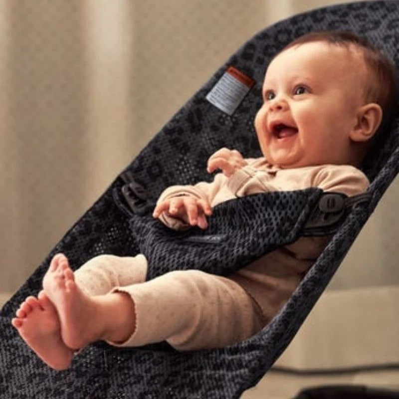 BabyBjorn Bouncer Bliss in dark patterned fabric with BABYBJORN tag; smiling baby reclines securely in the rocker.