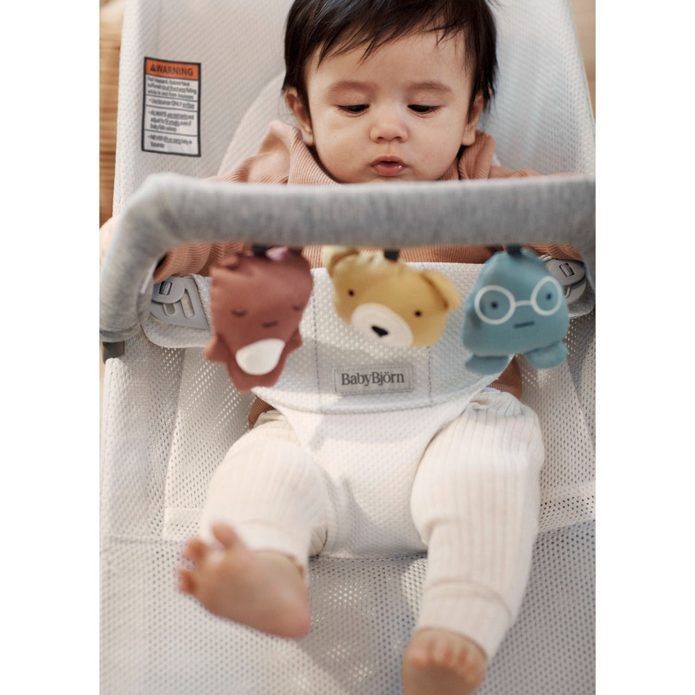 BabyBjorn Bouncer Toy in light gray mesh; baby sits and reaches for three hanging plush toys.