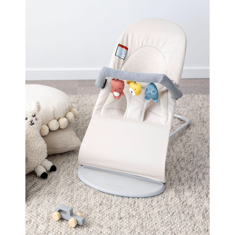 BabyBjorn Bouncer Toy in beige with a gray toy-bar and three hanging plush characters on a soft rug scene.