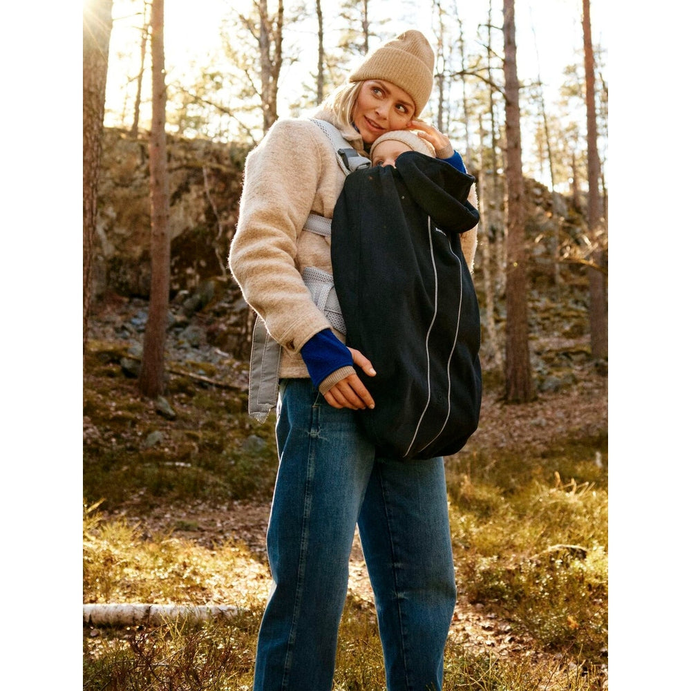 Mother outdoors in a forest, wearing a beige beanie, carries baby in a black BABY BJORN Cover for Baby Carrier.