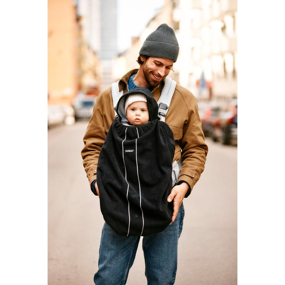 BabyBjorn Cover for Baby Carrier worn by a smiling dad carrying a baby on a city street in a black cover with white piping