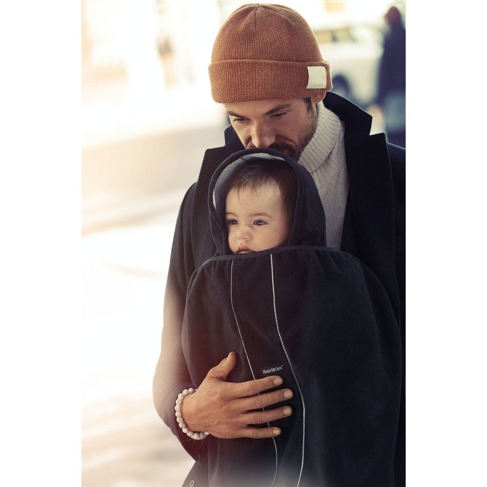 BABY BJORN Cover for Baby Carrier, navy; parent cradles baby in a warm, hooded carrier for cozy protection.