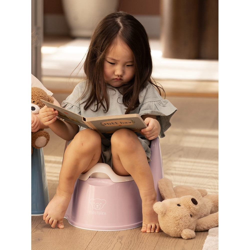 BabyBjorn Potty Chair in soft pink, a young girl sits reading a book on it with a teddy nearby.