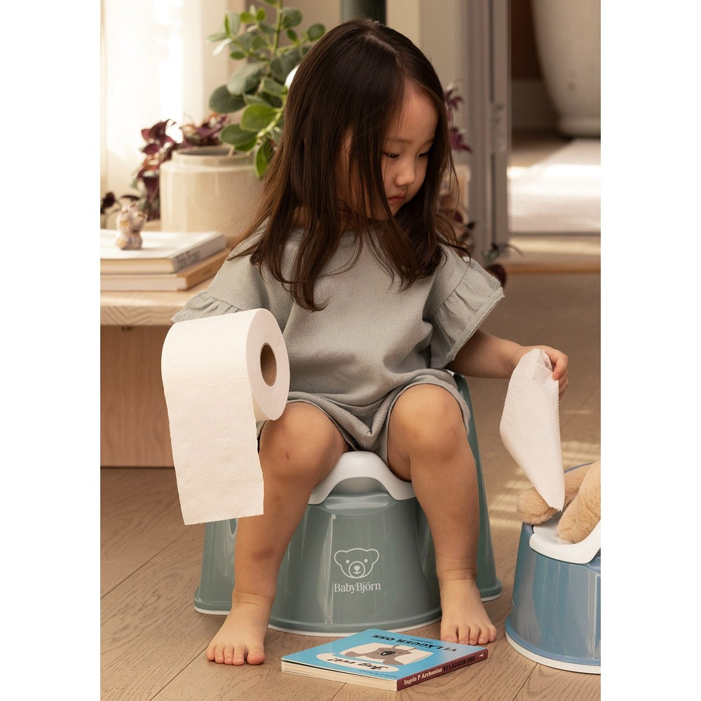 BabyBjorn Potty Chair in blue-green, toddler sits on it with a roll of toilet paper, practicing potty training.