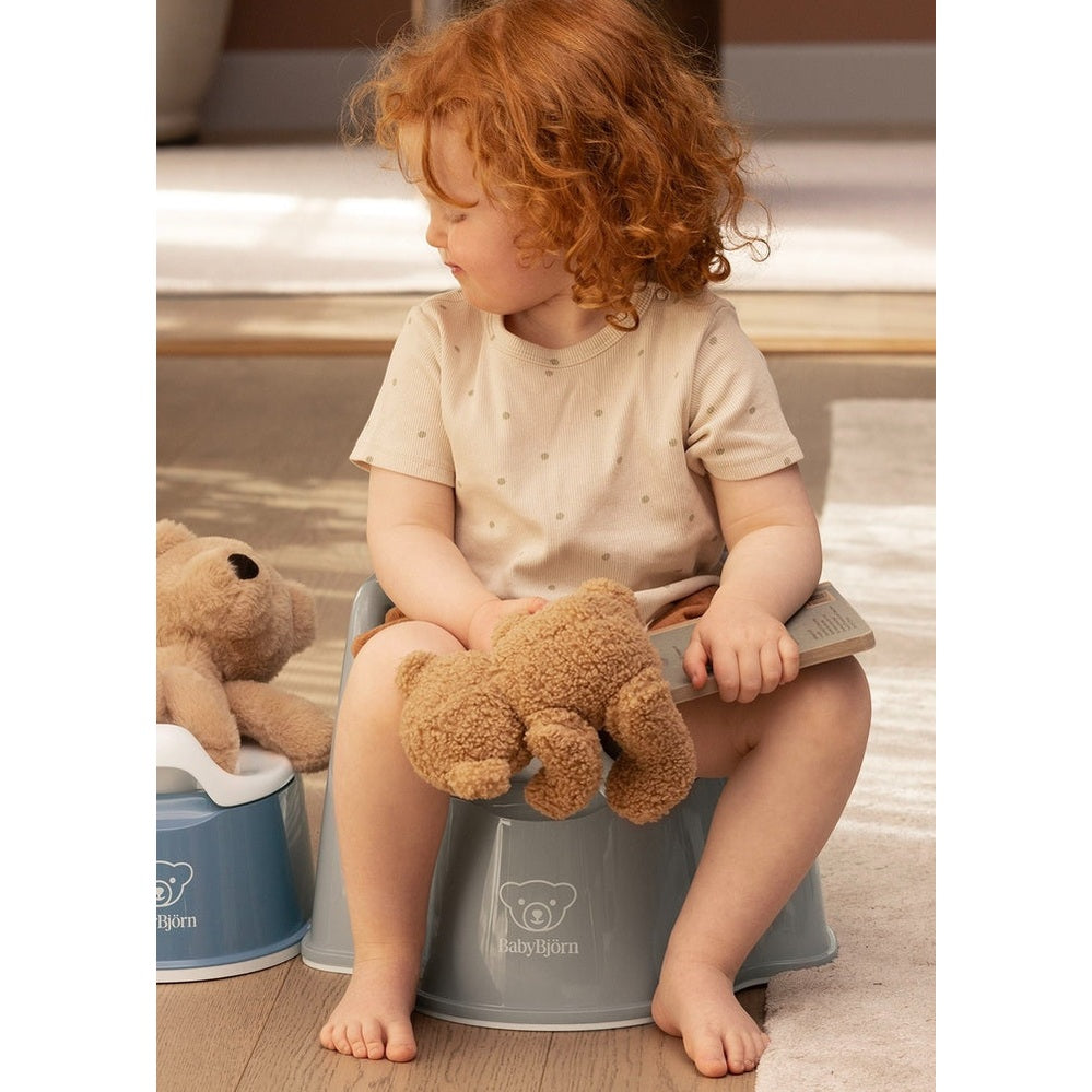 BabyBjorn Potty Chair gray with bear logo; toddler sits on it, holding a plush bear during potty training.