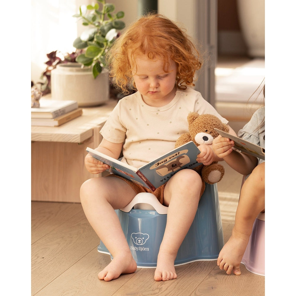 Toddler sits on BABYBJORN Potty Chair in blue, reading a book with a teddy bear in a sunny home.