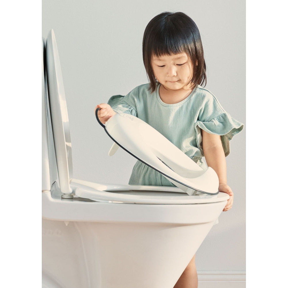 BabyBjorn Toilet Trainer: a white training seat being lifted onto the toilet by a young girl, pale-green dress.