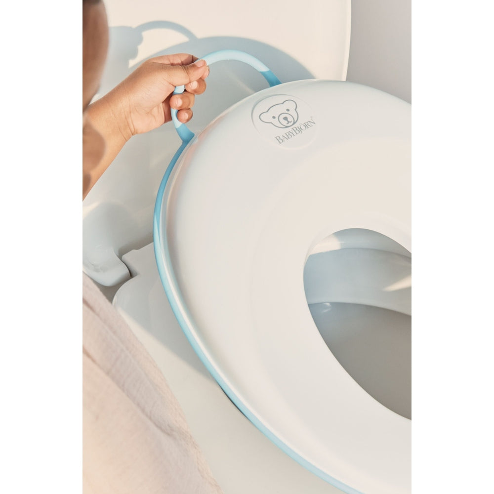 BabyBjorn Toilet Trainer in white with blue trim, where a child grips the side handle to steady it.