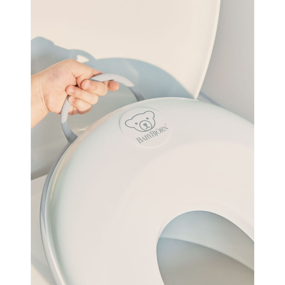 BabyBjorn Toilet Trainer in white with gray handle, hand carrying it to show portability.