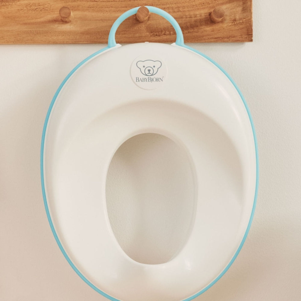 BabyBjorn Toilet Trainer in white with light-blue rim, hanging on wall for potty training.