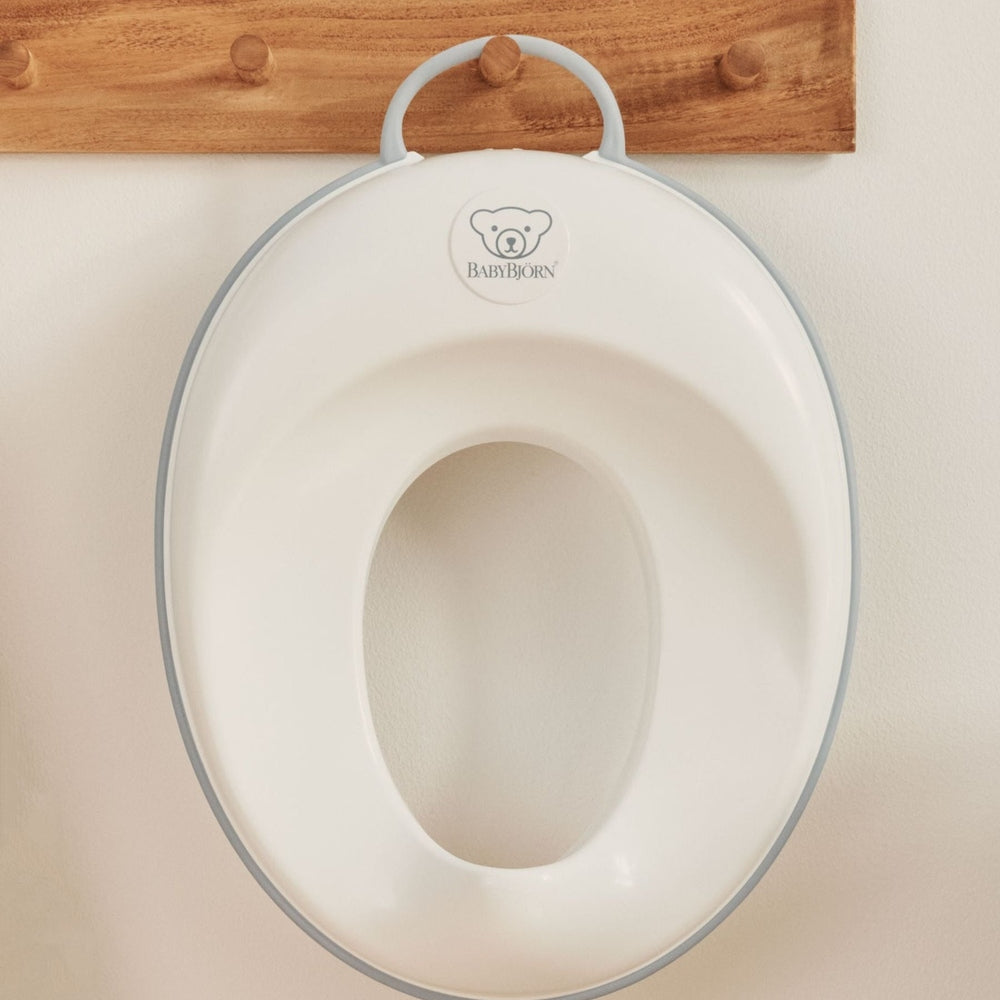 BabyBjorn Toilet Trainer in white with gray trim, hanging on a wooden wall hook.