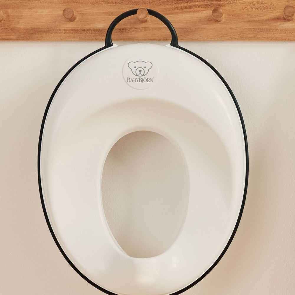 BabyBjorn Toilet Trainer, white with black edge, hanging on a wooden rack in a nursery.