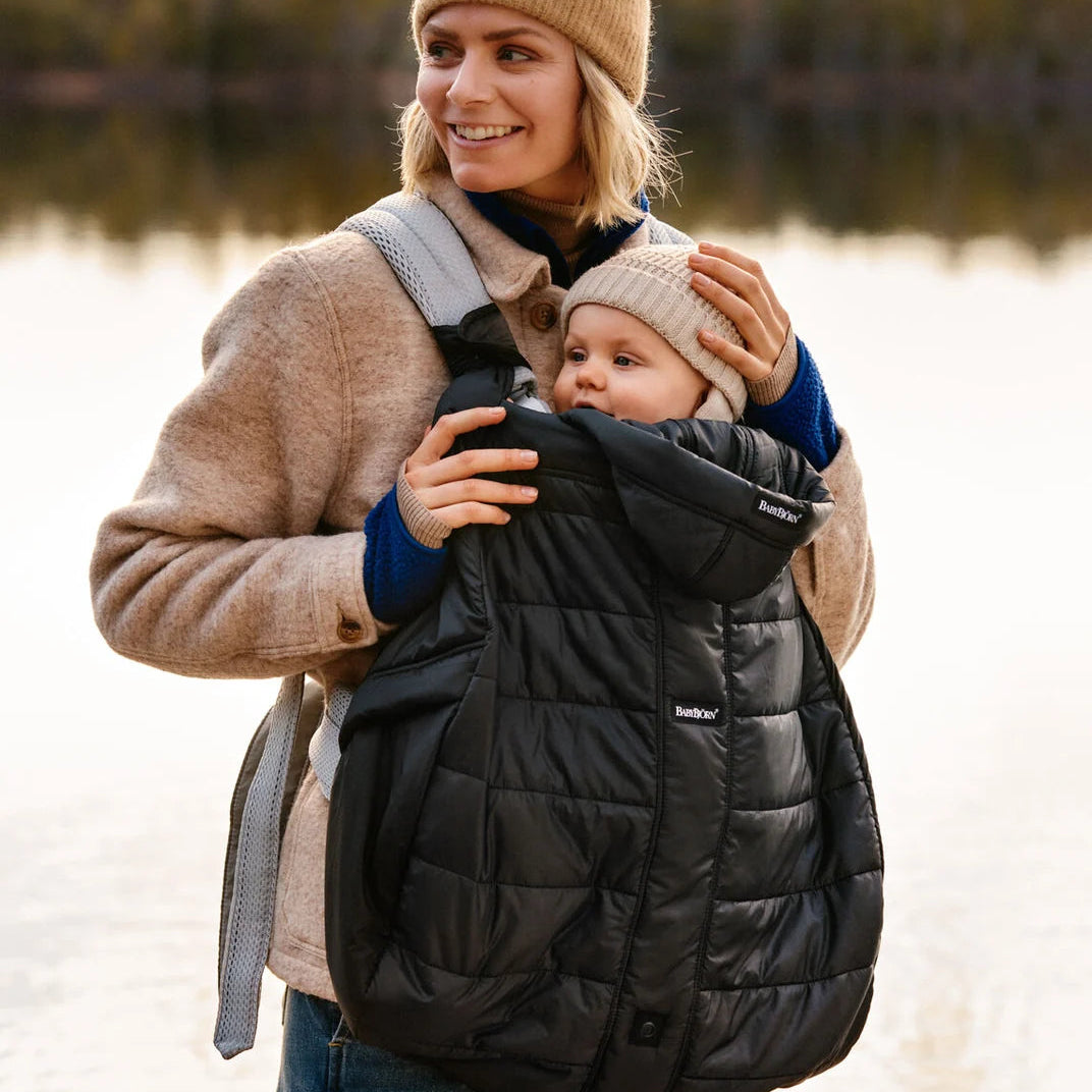 Parent carrying baby by a lake with BabyBjörn Winter Cover in Black