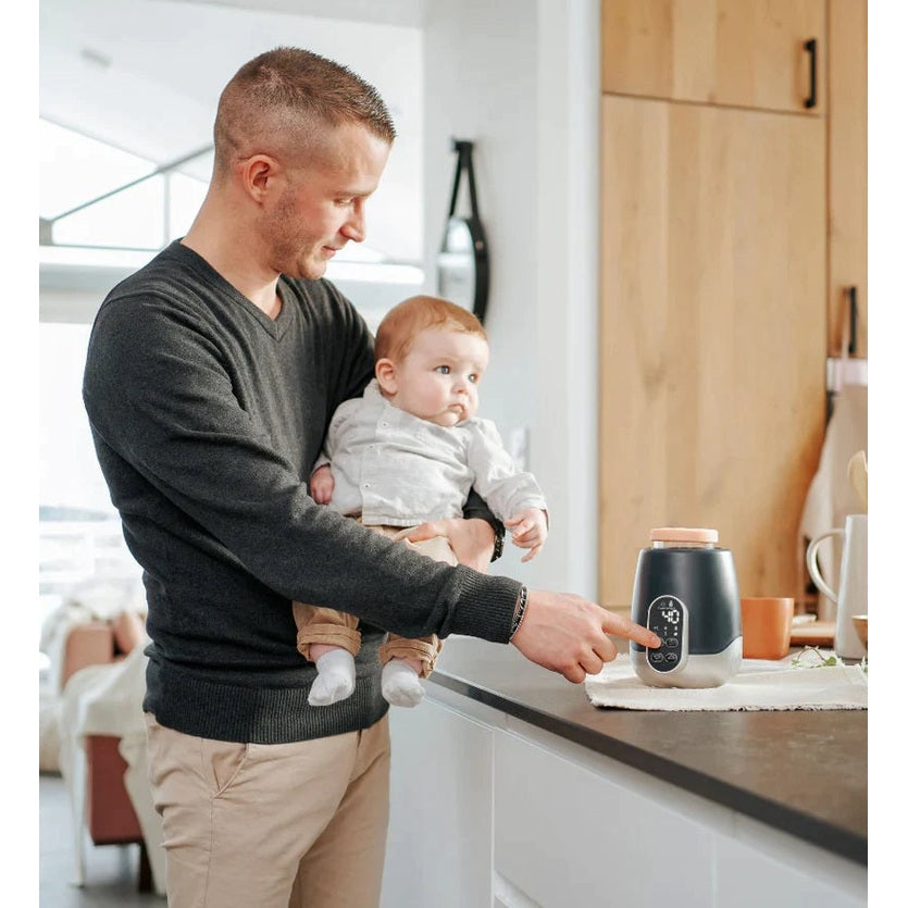 Babymoov Duo Smart Bottle Warmer with digital display on kitchen counter, dad holding baby.
