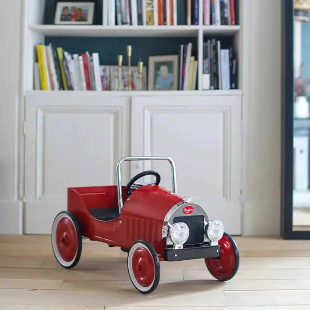 Baghera Ride-On Classic Pedal Car in red rests on a wooden floor near a bookshelf.