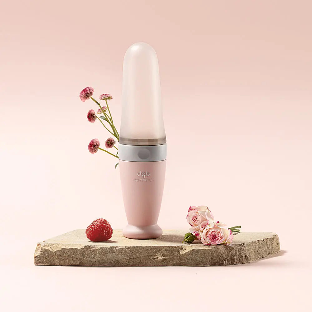 Bc Babycare Feeding Spoon Dispenser in soft pink with white band, upright on a stone platform beside pink flowers.