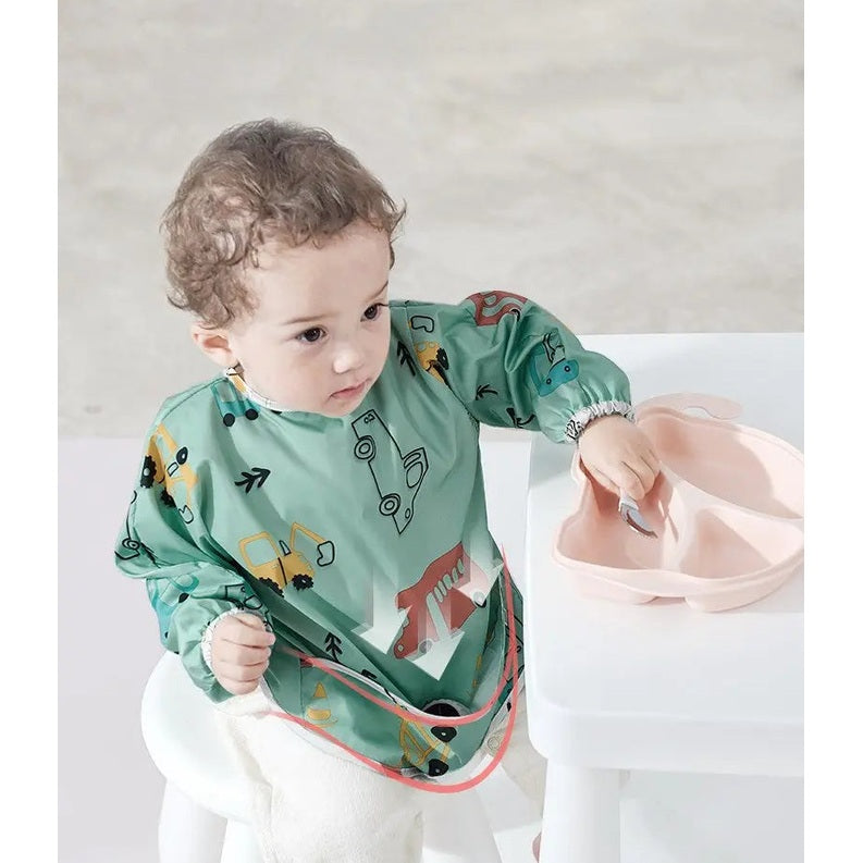 BC Babycare Long Sleeve Bib in green car print with front pocket, toddler wears it at table to catch spills.