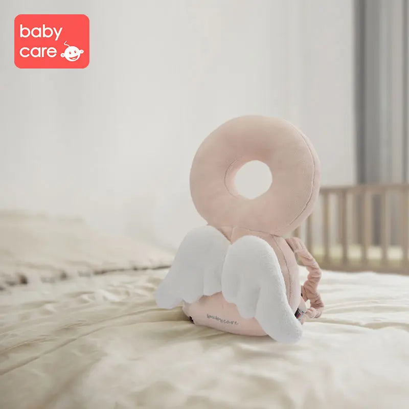 BC Babycare Protective Cushion plush neck pillow in pastel pink with white angel wings resting on a bed.