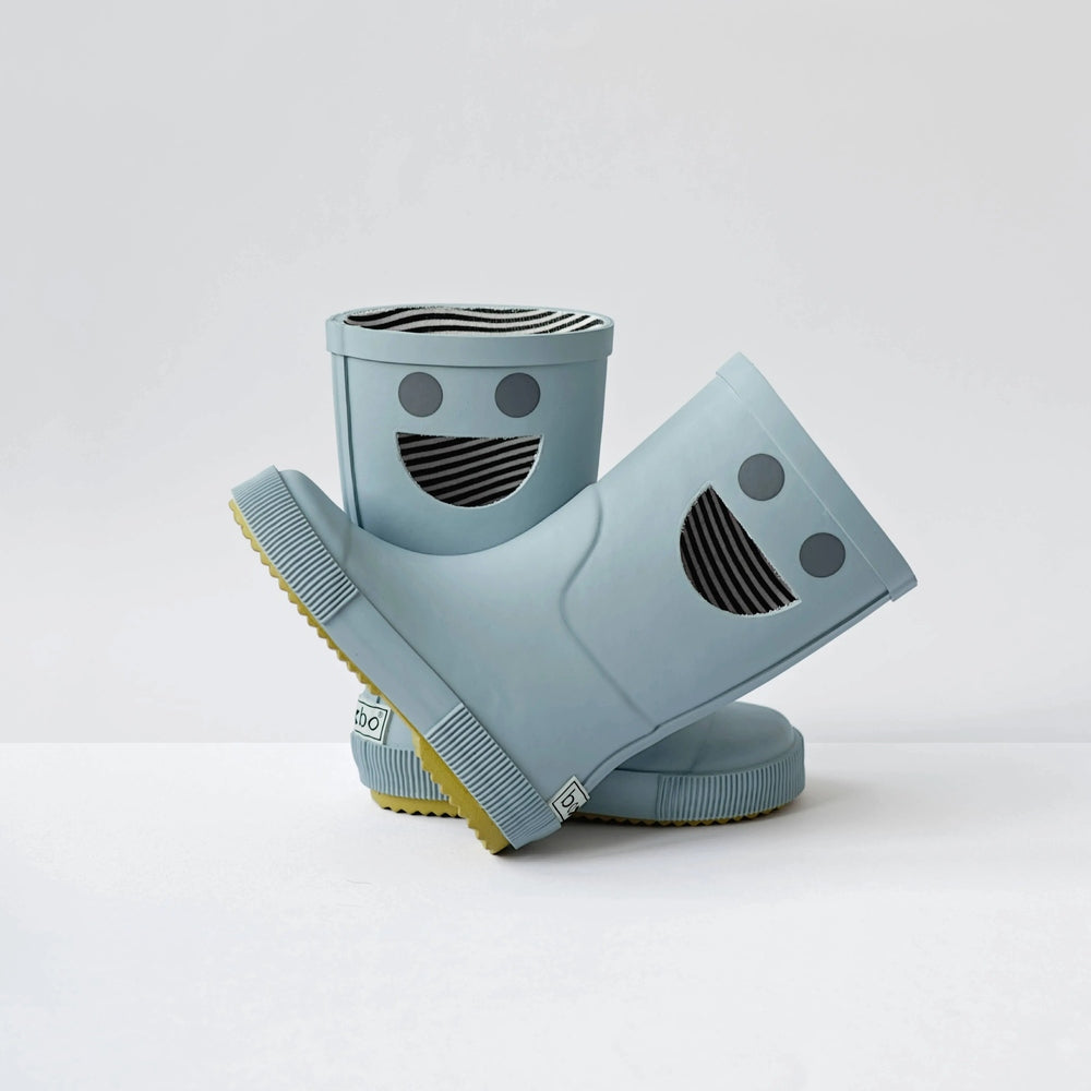 BOXBO Wistiti Rainboots Platine: light blue rubber boots with smiley cutouts, two shown, one leaning to reveal striped interior.