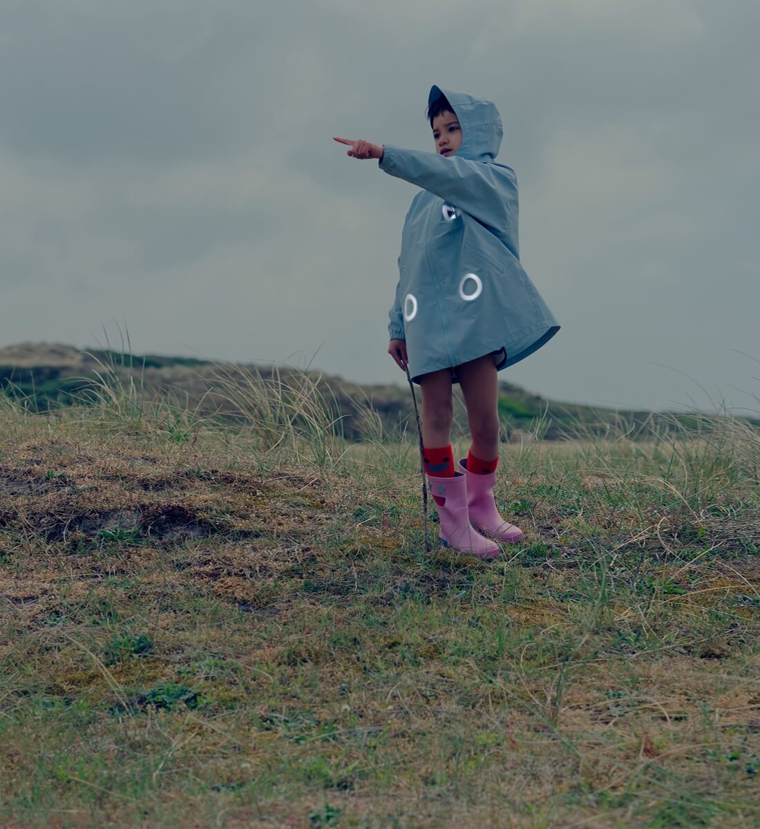 Boxbo Wistiti Windbreaker Rain Jacket Blue worn by a child outdoors with hood up, pointing in a windy field.