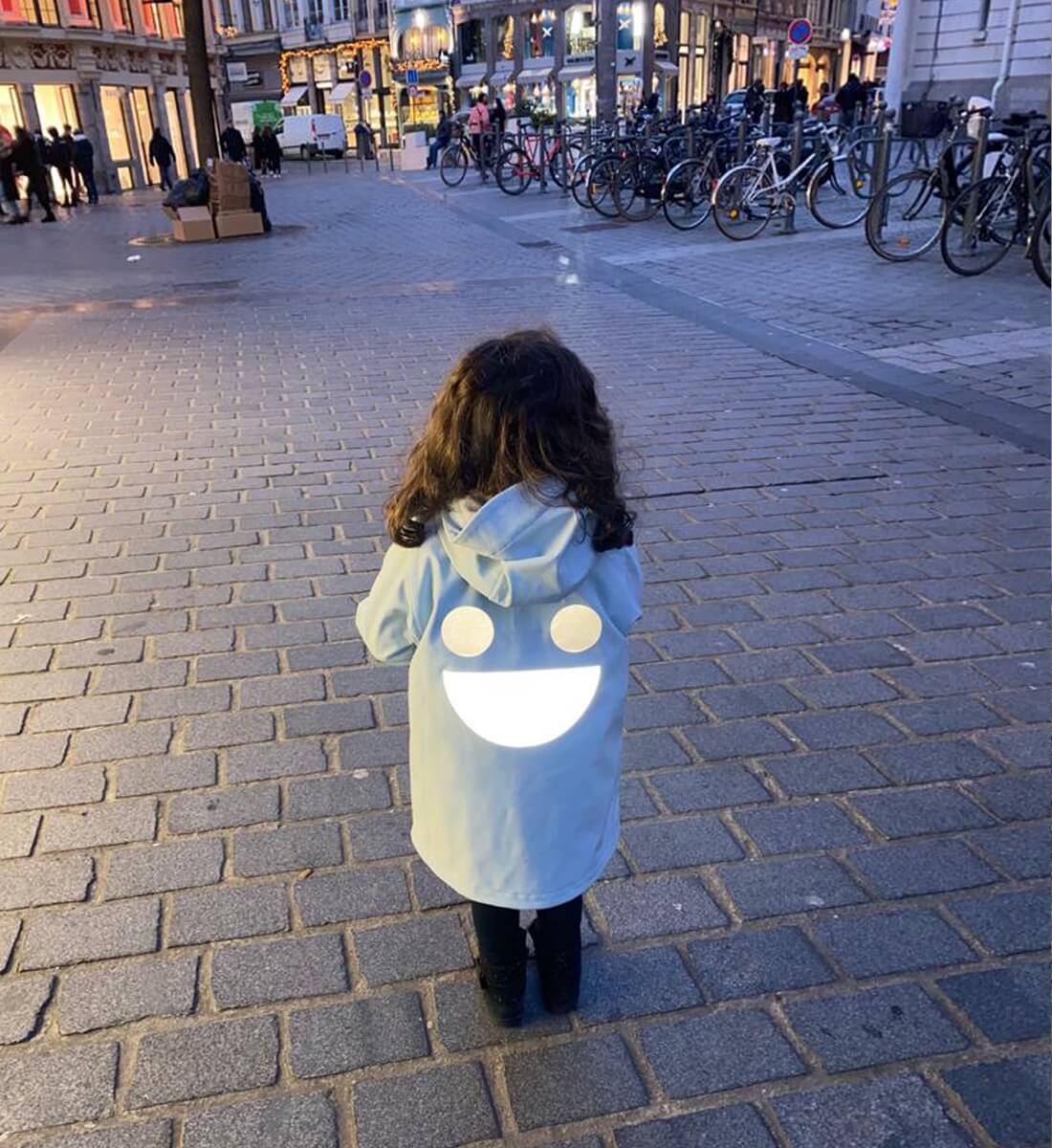 Boxbo Wistiti Windbreaker Rain Jacket Blue, hooded child walking on a city street at dusk with a large white back smile.