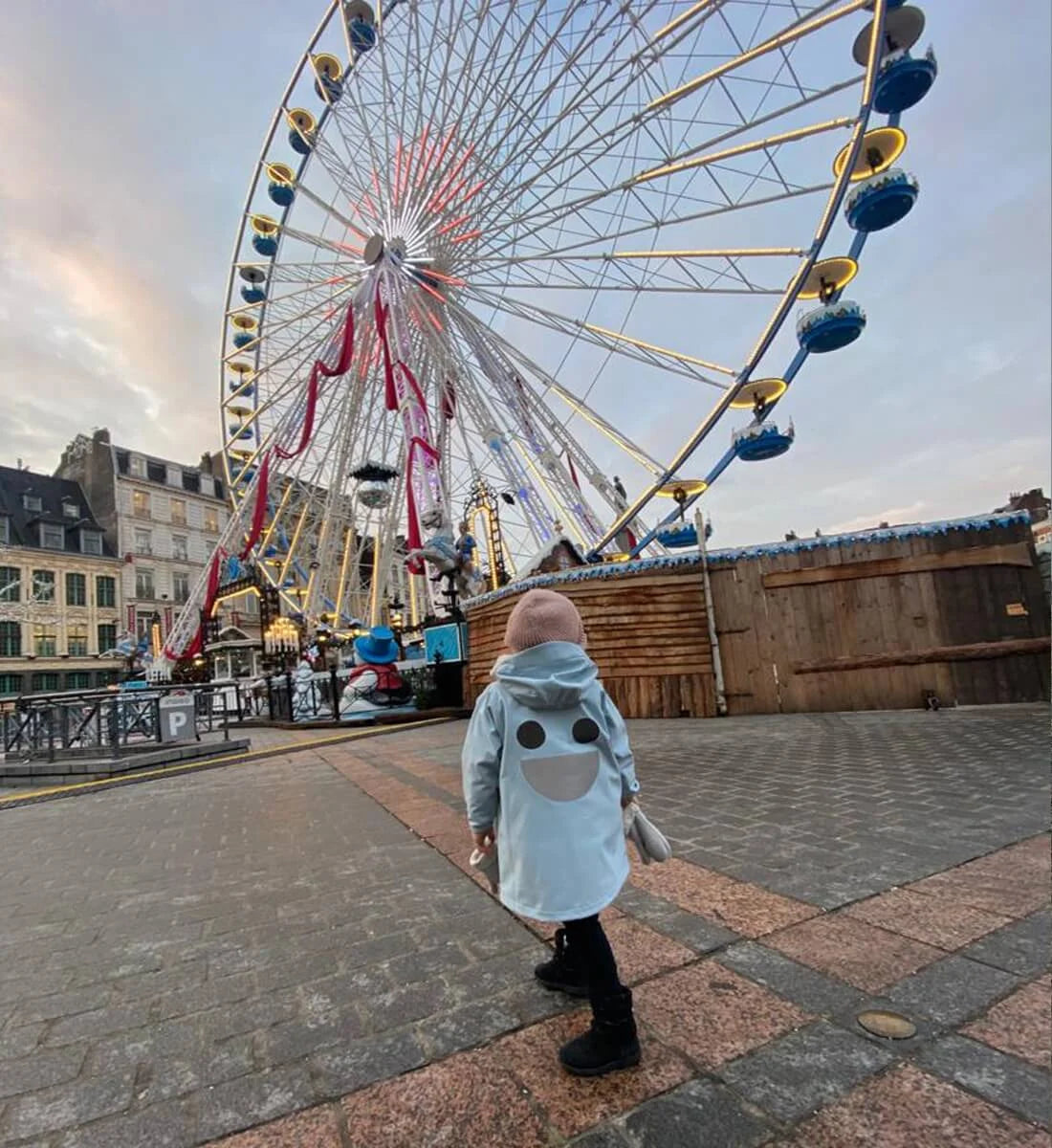 Child wearing a blue BOXBO Wistiti rain jacket near a Ferris wheel