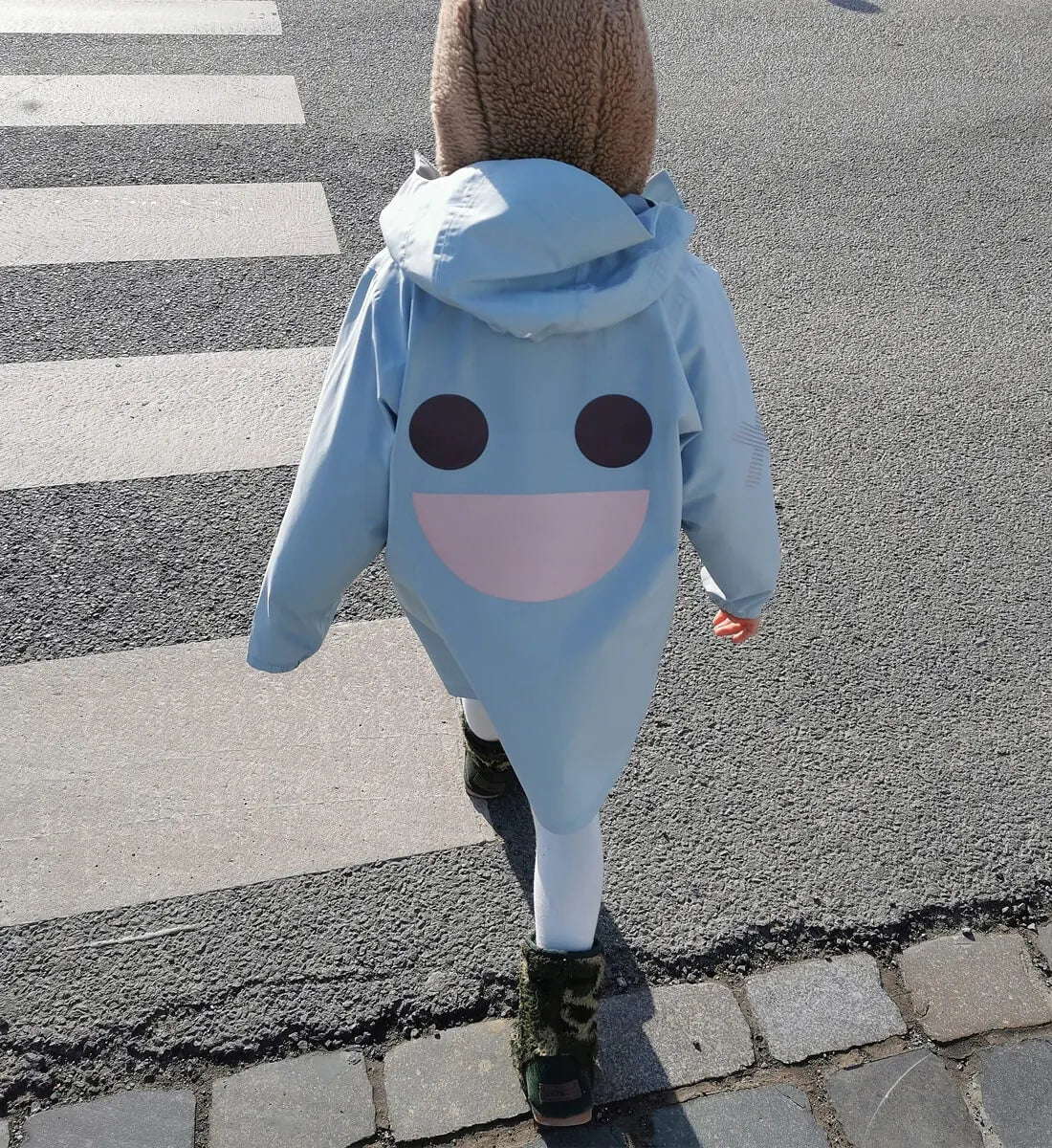 Child wearing a blue BOXBO Wistiti rain jacket while crossing a street, back view