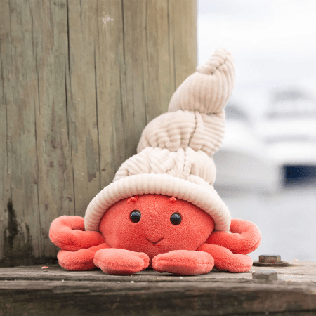 Bukowski Bears Hermit The Crab stuffed animal with coral body and cream knitted shell, resting on a wooden dock.