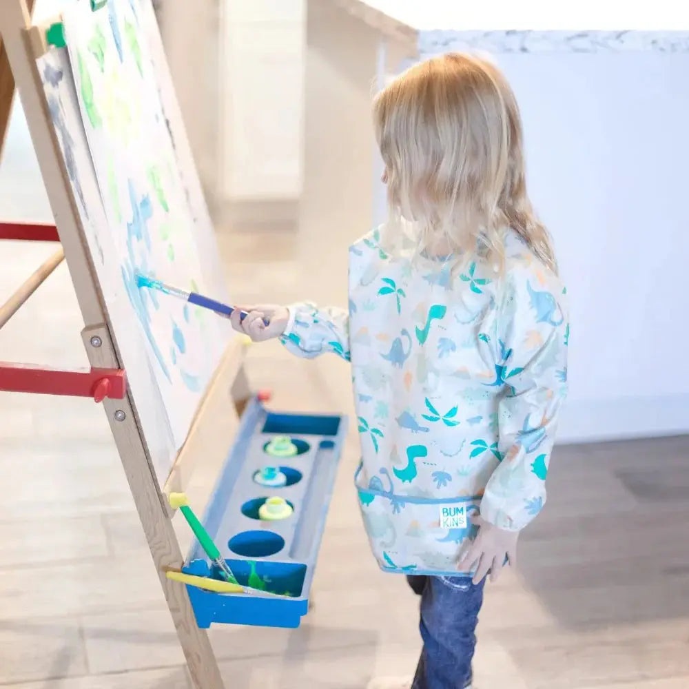 Bumkins Long Sleeved Smock Bib in blue dinosaur print, a child paints at an easel to keep clothes clean.