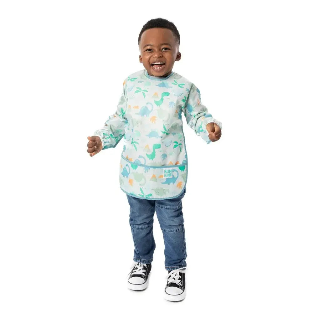 Bumkins Long Sleeved Smock Bib in pastel dinosaur print with front pocket, worn by a smiling toddler.