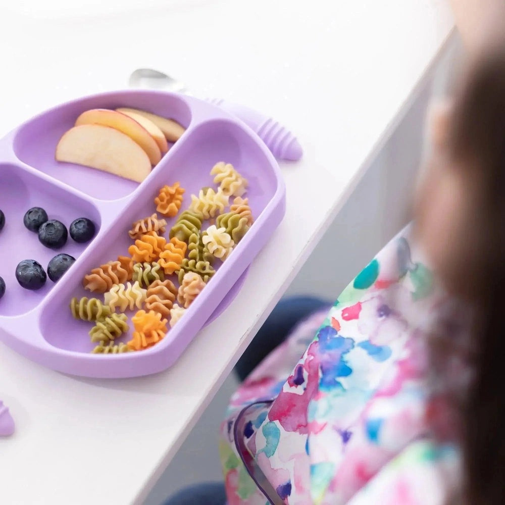 Bumkins Silicone Grip Dish in purple, divided sections with blueberries, apple slices, and colorful pasta for baby feeding.