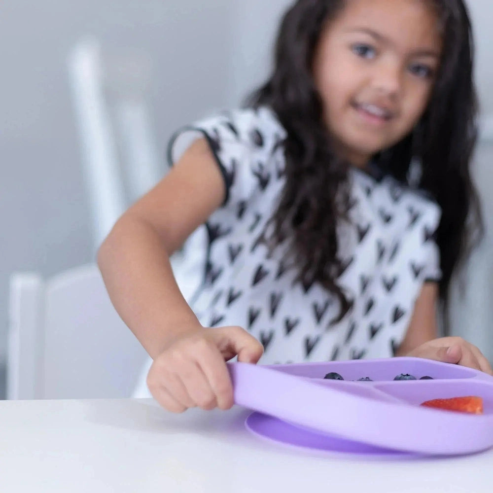 Bumkins Silicone Grip Dish, purple, suction base shown as a child lifts it during mealtime.