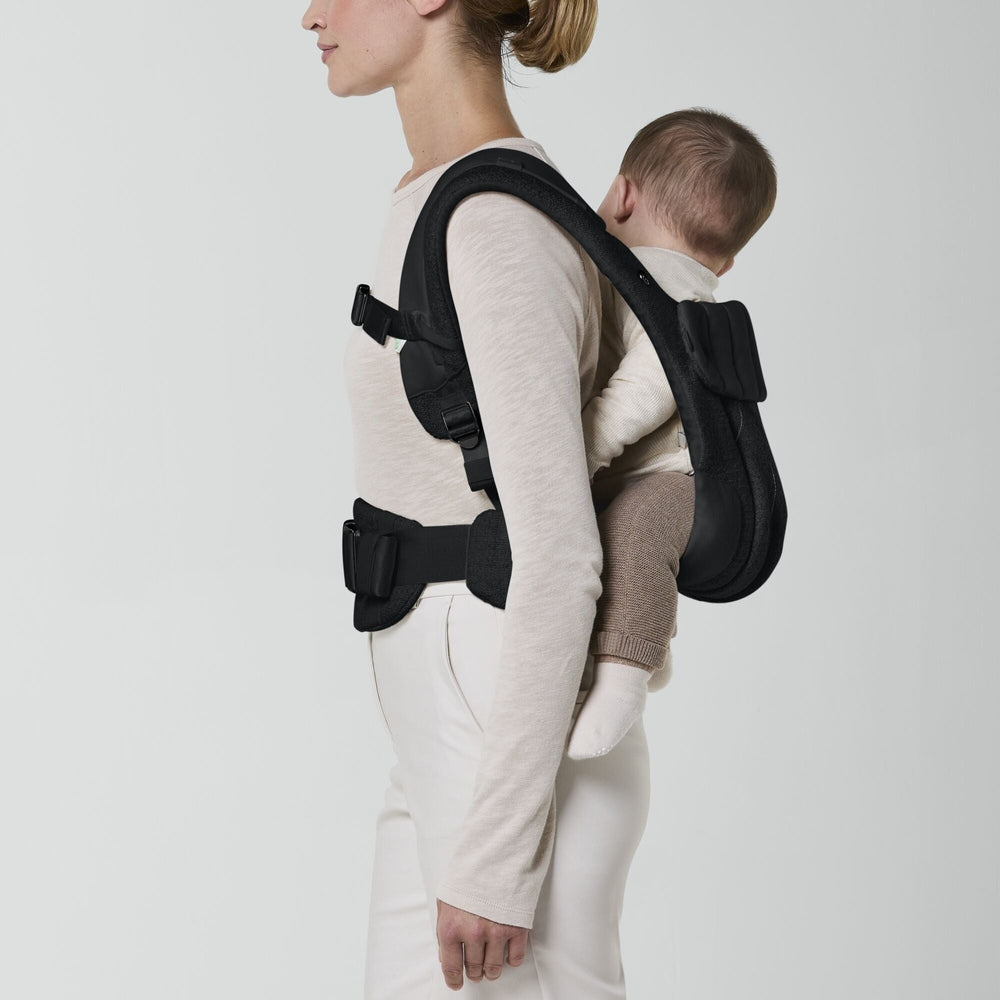 Close-up of the Cybex Coya Carrier Boucle black boucle cybex coya baby carrier worn by adult in beige top; infant seated on back with padded shoulder straps and waist belt.
