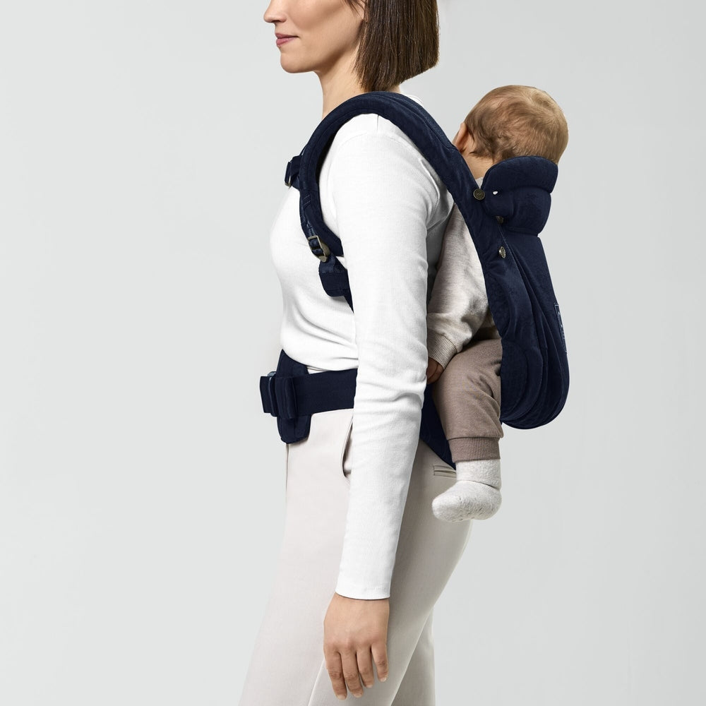Close-up of the Cybex Coya Carrier Rebellious Luxury Denim Blue navy denim-blue baby carrier worn by a person in white; padded shoulder straps and waist belt, baby seated on back.