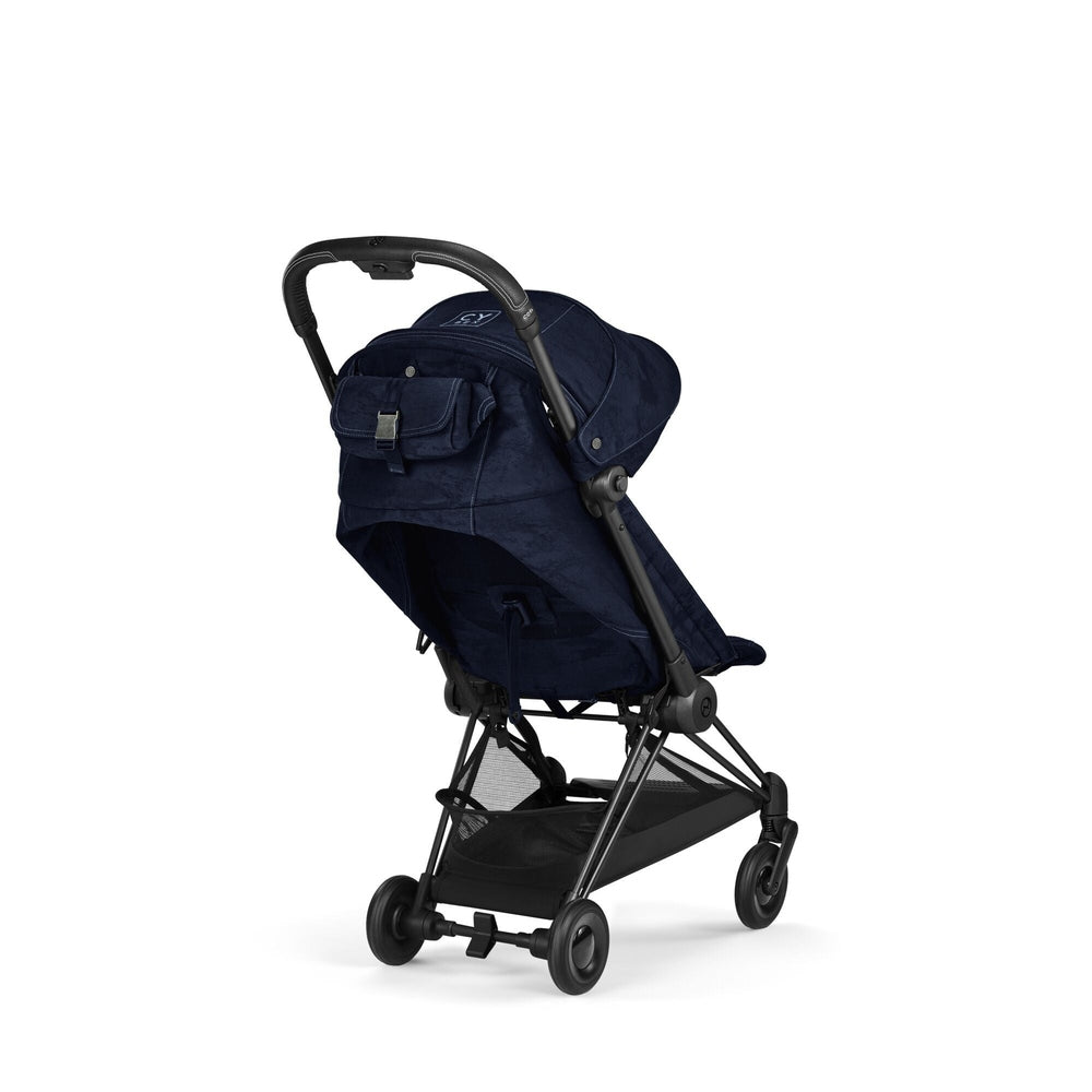 Close-up of the Cybex Platinum Coya Compact Lightweight Stroller - Rebellious Luxury Denim Blue denim blue fabric compact lightweight stroller with black frame, canopy, and mesh storage basket.