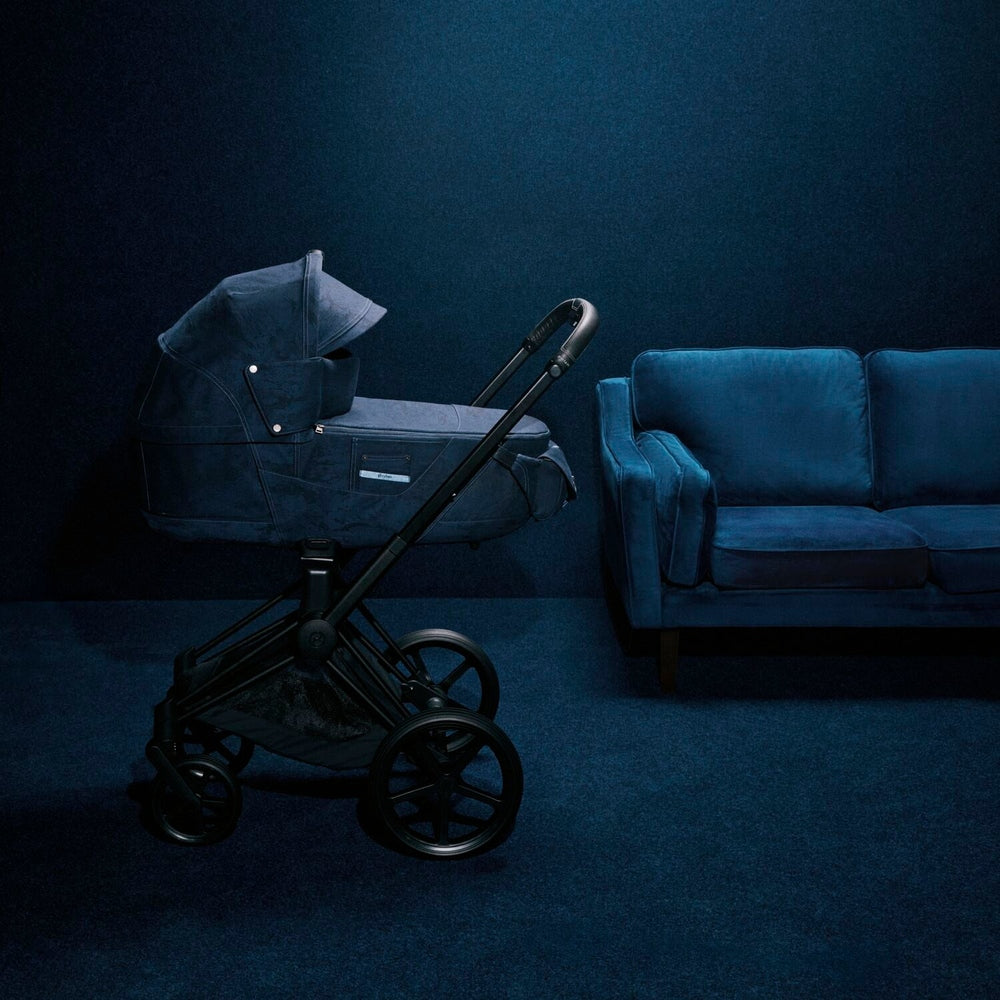 Close-up of the Cybex Platinum Priam Carry Cot Limited Edition - Rebellious Luxury Denim Blue denim-blue cybex priam carry cot on black chassis with large wheels beside a blue velvet sofa.