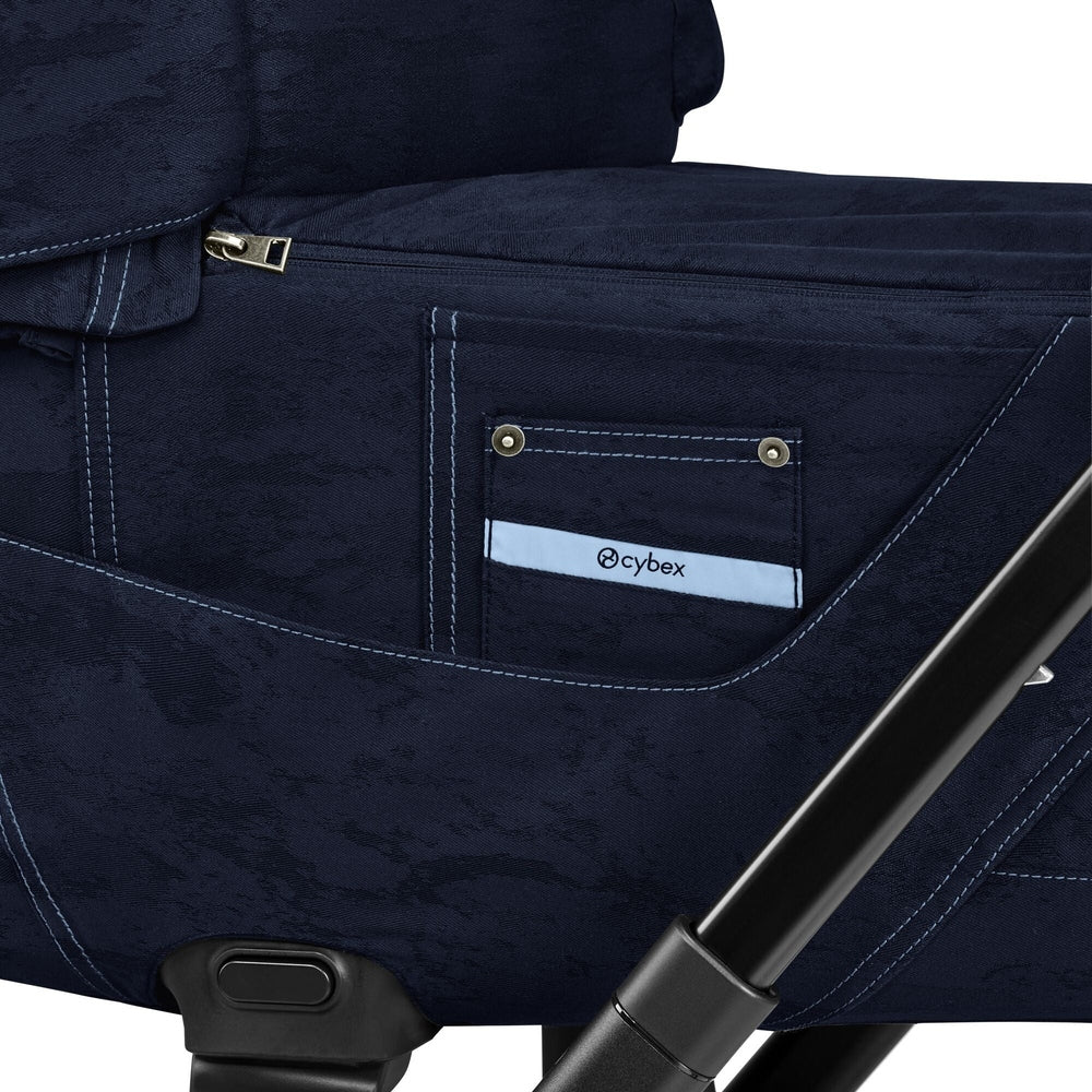 Close-up of the Cybex Platinum Priam Carry Cot Limited Edition Rebellious Luxury Denim Blue in textured navy fabric with contrast stitching, side pocket, zipper detail, and black frame bar.