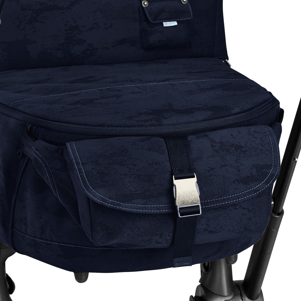 Close-up of the Cybex Platinum Priam Carry Cot Limited Edition - Rebellious Luxury Denim Blue with front pocket and silver buckle on a dark stroller frame.