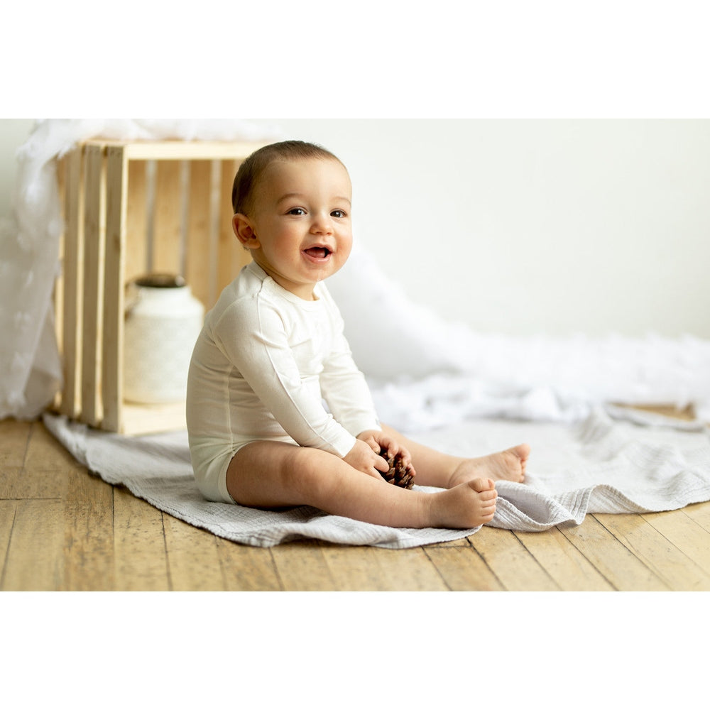 Coccoli Infant Cotton TENCELTM Modal Long Sleeve Bodysuit Cream, baby sits on a blanket holding a pinecone.