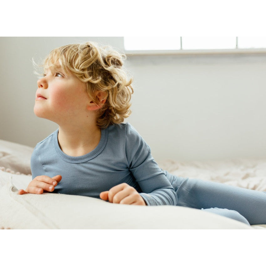 Coccoli Kid Long Sleeve Cotton TENCELTM Modal Pyjamas in steel blue jersey, a child lounging on a bed looking up.