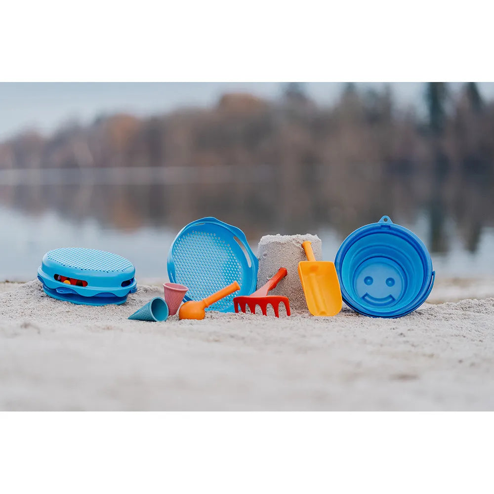 Colorful sand toys including buckets and shovels on a sandy beach with water in the background.