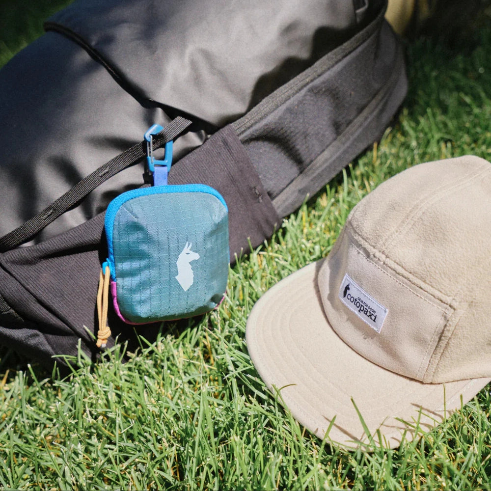 Cotopaxi Allpita Mini Bag in Teal with blue zipper, tan paracord pull, white llama logo, attached to black backpack.