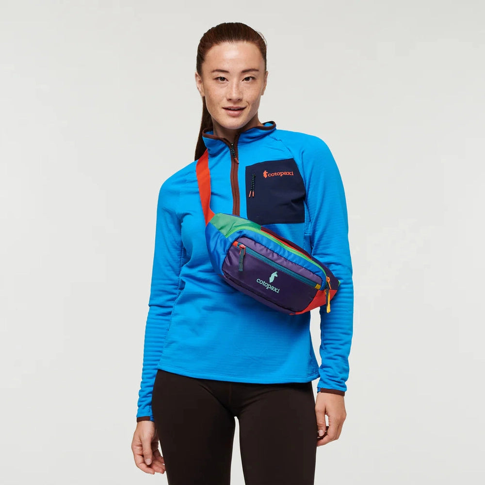 Cotopaxi Kapai 3L Hip Pack in purple with teal zippers, orange strap worn crossbody over a bright blue top