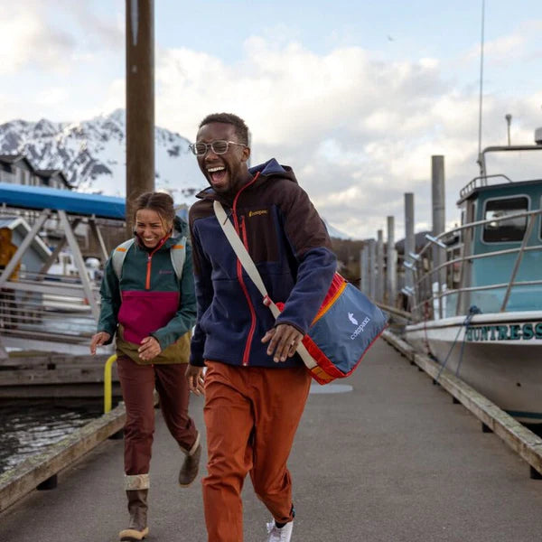 Cotopaxi Taal Convertible Tote in blue with orange interior worn crossbody on man at marina